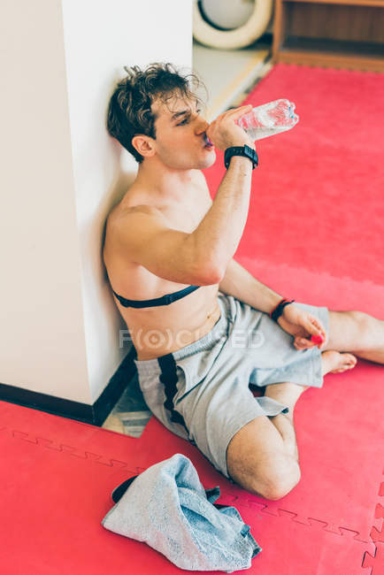 Hombre sentado apoyado en la columna con monitor de frecuencia cardíaca en el pecho, bebiendo agua de la botella - foto de stock
