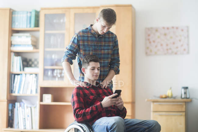 Молодий чоловік використовує інвалідне крісло для читання текстів смартфона на кухні — стокове фото