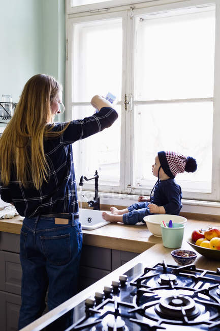 Femme adulte moyenne versant de l'eau dans l'évier de cuisine pour bébé fils — Photo de stock