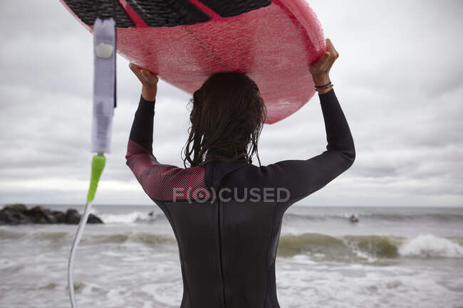 Vista trasera de la surfista femenina llevando tabla de surf en la cabeza en Rockaway Beach, Nueva York, EE.UU. - foto de stock