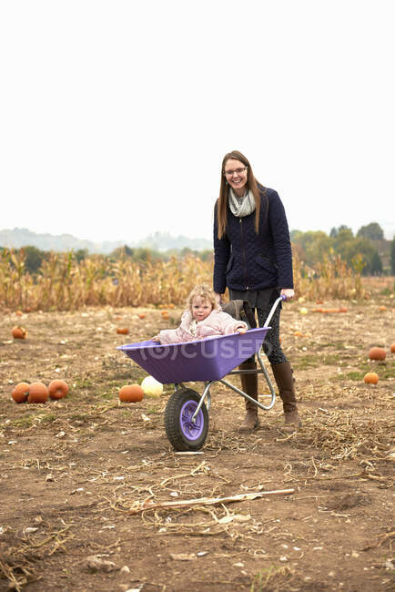 Portrait of mid adult woman pushing toddler daughter in wheelbarrow in pumpkin field — Stock Photo
