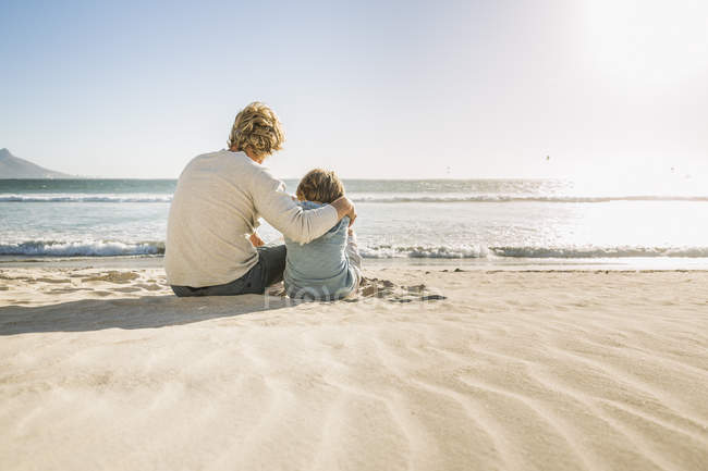 Padre e hijo sentados en la playa mirando hacia otro lado - foto de stock
