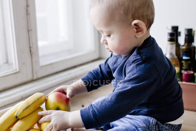 Baby boy sitting on kitchen counter playing with bowl of fruit — Stock Photo