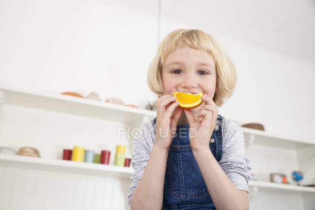 Retrato de bonito menina no cozinha segurando laranja fatia para ela boca — Fotografia de Stock