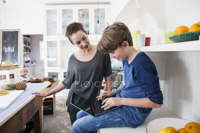 Mujer joven y niño en la cocina, niño sentado en la superficie de trabajo utilizando el ordenador portátil - foto de stock