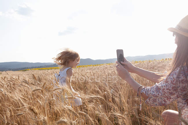 Mother photographing girl running through wheat field — Stock Photo