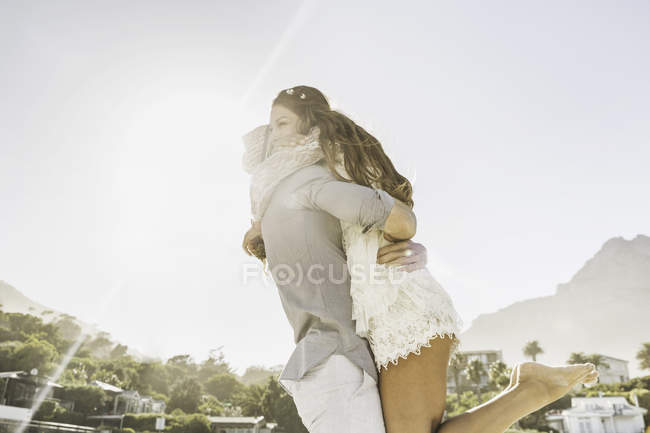 Uomo che solleva e abbraccia fidanzata sulla spiaggia illuminata dal sole, Città del Capo, Sud Africa — Foto stock