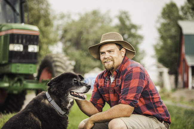 Agricultor vistiendo sombrero de vaquero acariciando perro en la granja mirando hacia otro lado sonriendo - foto de stock