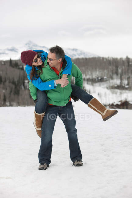 Reifer Mann trägt junge Frau auf dem Rücken im Schnee — Stockfoto