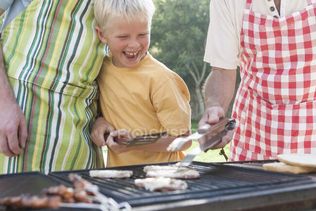 Boy cooking kebabs and burgers on barbecue with father and grandfather — Stock Photo