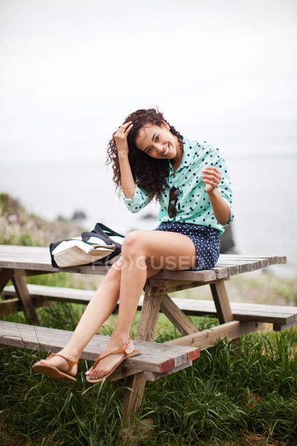 Jeune femme assise sur une table de pique-nique souriant, portrait — Photo de stock