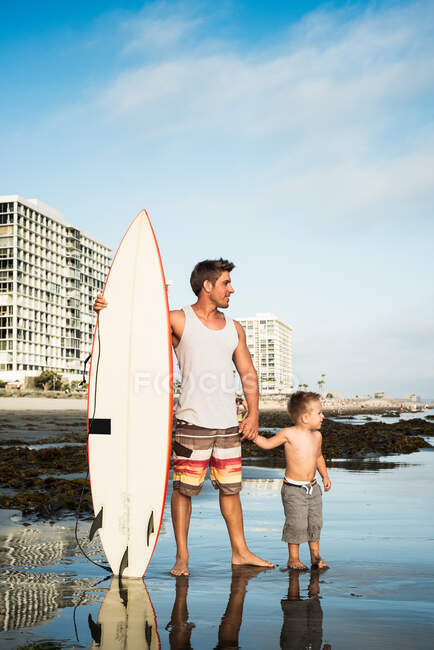 Jovem segurando prancha de surf com filho no mar — Fotografia de Stock
