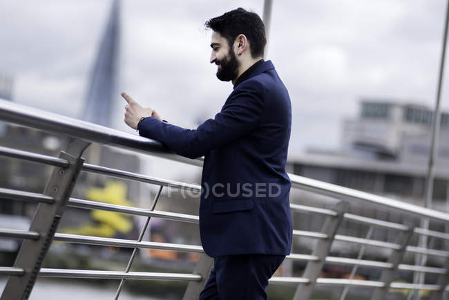 Бизнесмен, стоящий на пешеходном мосту и читающий текст смартфона, Лондон, Великобритания — стоковое фото