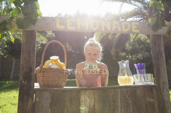 Porträt eines Mädchens auf Limonadenständer, das einen Dollar-Schein hochhält — Stockfoto