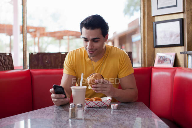 Hombre joven mensajes de texto en el teléfono móvil y comer comida rápida en la cafetería - foto de stock