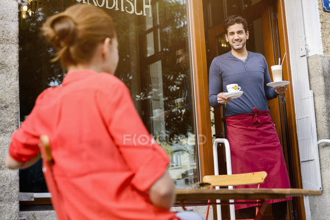 Giovane donna seduta fuori caffè, operaio caffè portando bevande fuori — Foto stock