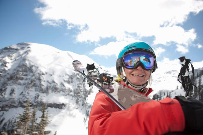 Junge Frau in Skibekleidung mit Skiern über der Schulter, lächelnd — Stockfoto