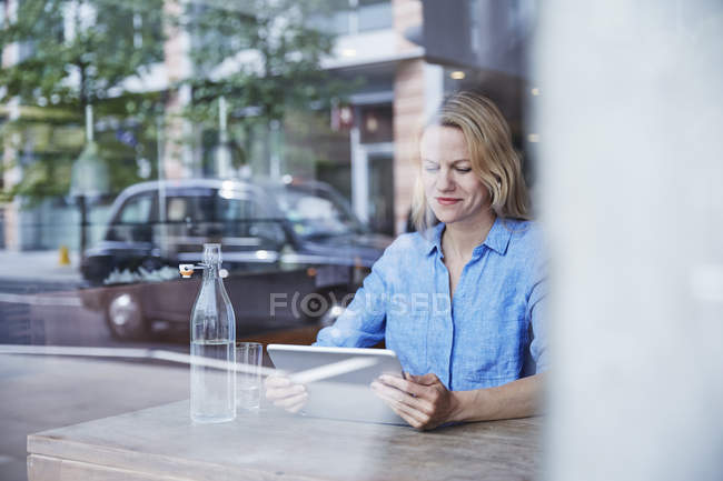 Reife Frau sitzt im Café, nutzt digitales Tablet, Taxi spiegelt sich im Fenster — Stockfoto