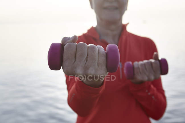 Mature woman beside water, exercising with hand weights, mid section — Stock Photo