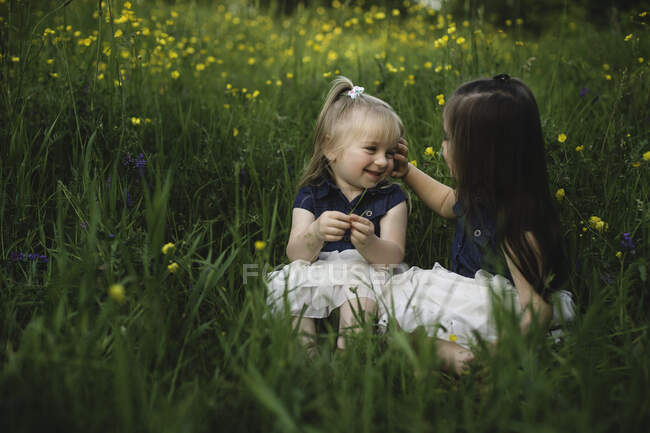 Chicas sentadas en el prado de flores silvestres cara a cara sonriendo - foto de stock