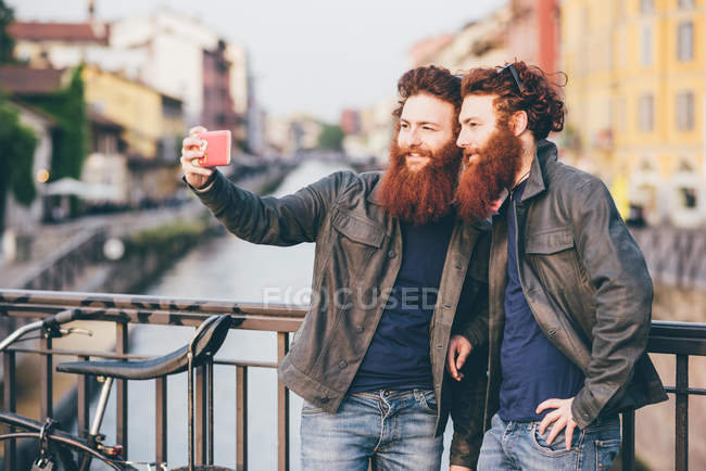 Молоді чоловіки хіпстер близнюки з рудим волоссям і бородою беруть смартфон селфі на набережній каналу — стокове фото