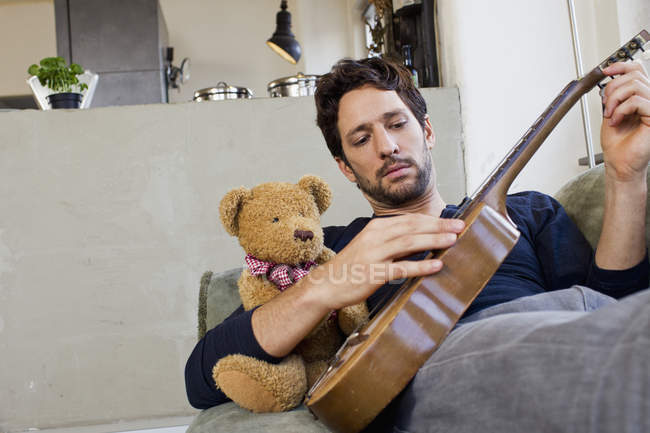 Середній дорослий чоловік сидить на дивані, граючи на гітарі з плюшевим ведмедем — стокове фото