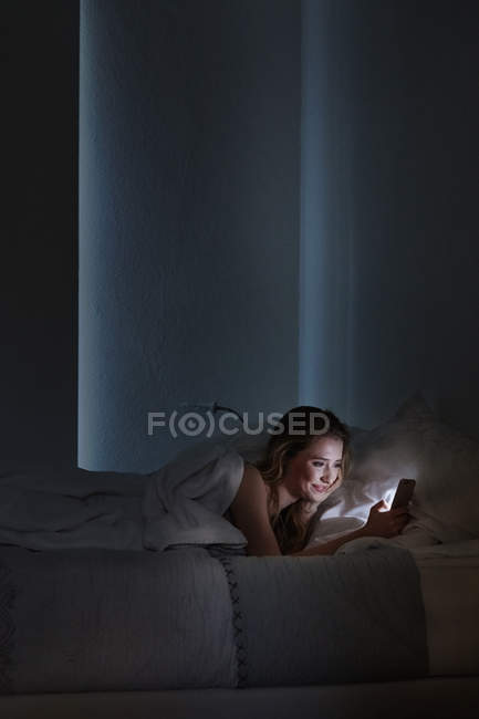 Jovem deitada na cama lendo textos de smartphones à noite — Fotografia de Stock