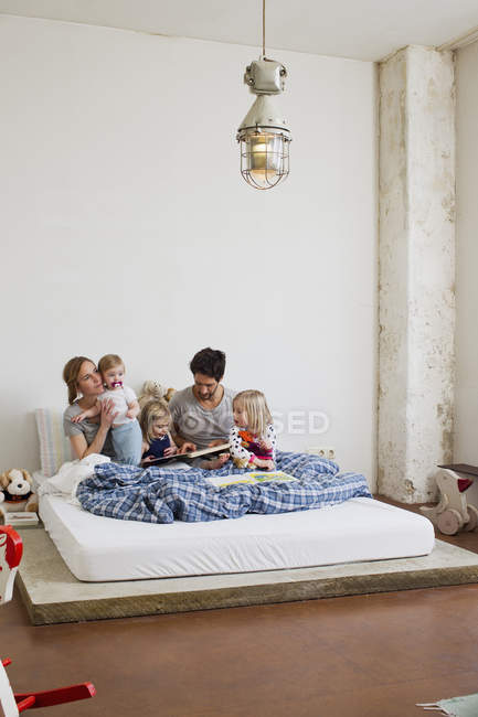 Семья с тремя дочерьми читает книги в постели — стоковое фото