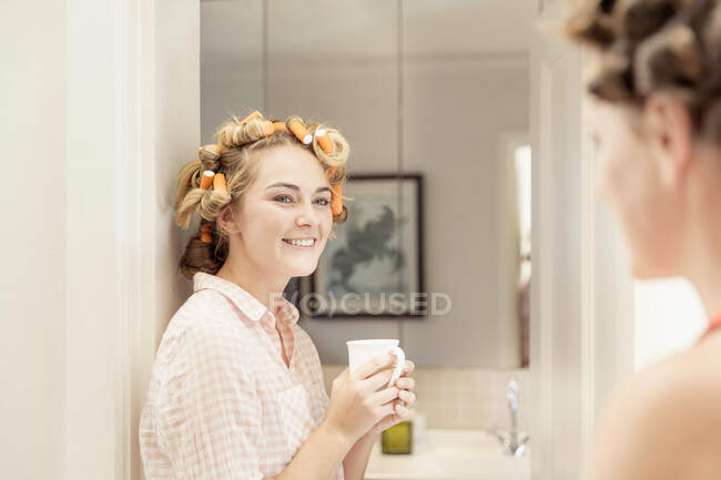 Jeune femme, rouleaux de mousse dans les cheveux, tenant une boisson chaude, souriant à un ami — Photo de stock