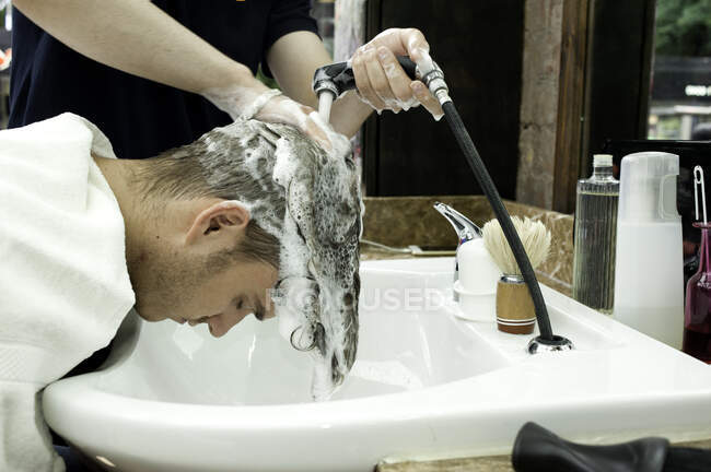 Vista lateral do jovem se curvando para a frente tendo o cabelo lavado na barbearia — Fotografia de Stock
