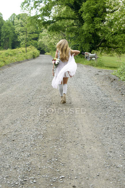 Vista trasera de una joven mujer caminando por un camino rural con flores a sus espaldas - foto de stock