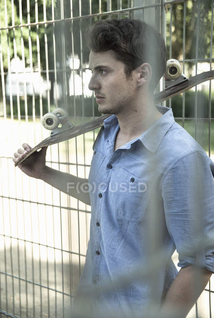 Serious young man with skateboard on shoulder behind park fence — Stock Photo