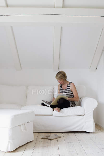 Зріла жінка сидить хрест ногами на дивані читає книгу — стокове фото