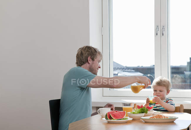 Padre e hijo pequeño desayunando en la mesa de la cocina - foto de stock