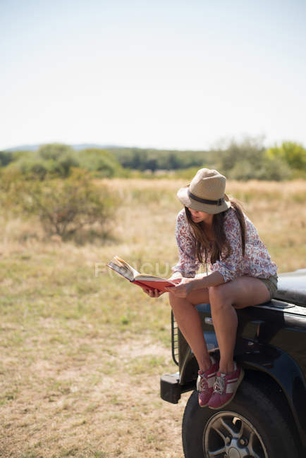 Mitte erwachsene Frau sitzt auf Motorhaube des Autos und liest Buch — Stockfoto