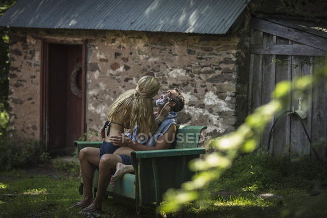 Jeune femme en bikini assis sur les genoux de petits amis se rasant le menton au chalet de vacances — Photo de stock