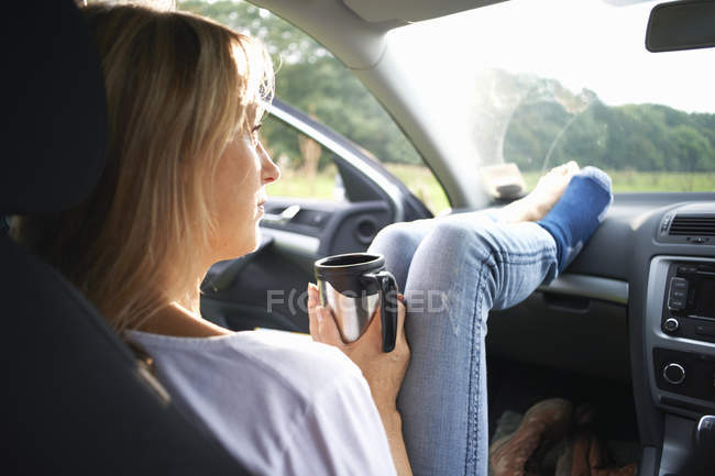 Жінка сидить в машині і тримає туристичну чашку кави — стокове фото