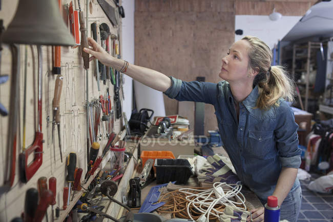 Женщина в мастерской тянется за инструментами, висящими на стене — стоковое фото