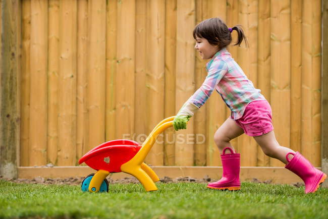Menina jovem correndo com carrinho de mão de brinquedo no jardim — Fotografia de Stock