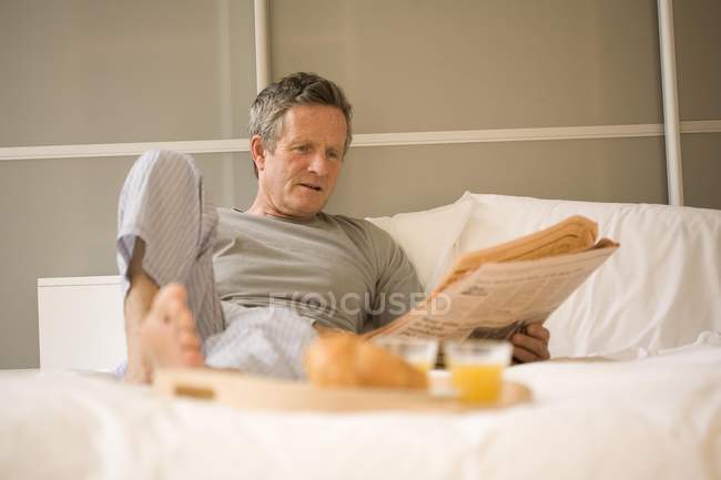 Senior man sitting up in bed reading newspaper — Stock Photo