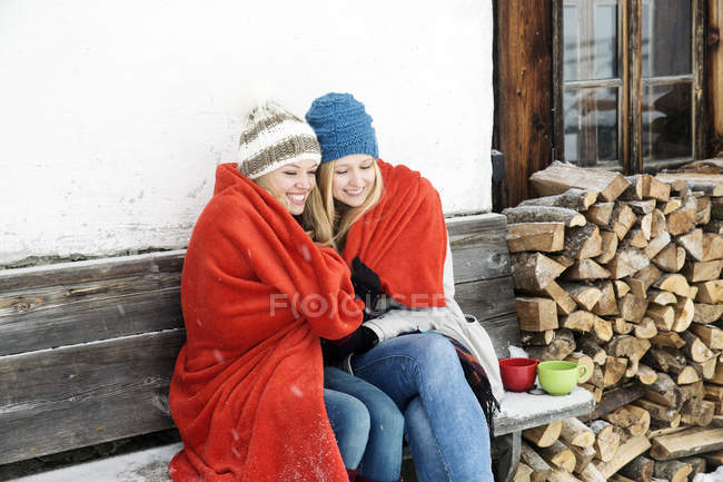 Двоє молодих друзів, загорнуті в червону ковдру, сидять зовні дерев'яної кабіни — стокове фото