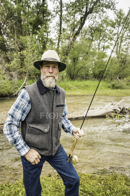 Homem barbudo por rio segurando haste de pesca olhando para a câmera — Fotografia de Stock