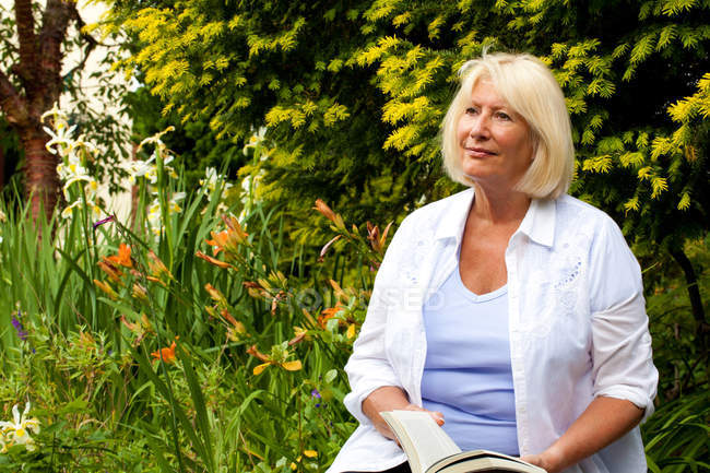 Mujer mayor sentada en el jardín sosteniendo libro - foto de stock