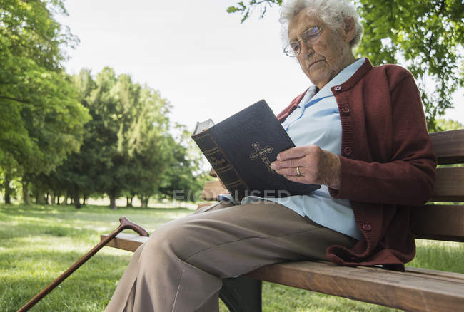 Mulher sênior sentada no banco do parque e lendo bíblia — Fotografia de Stock