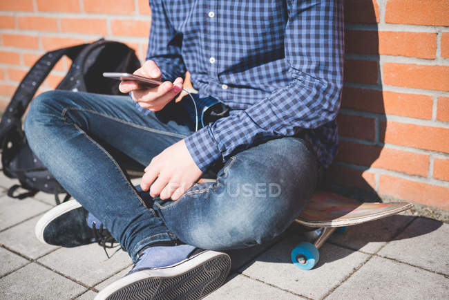 Neck down view of young male urban skateboarder sitting on sidewalk reading smartphone text — Stock Photo