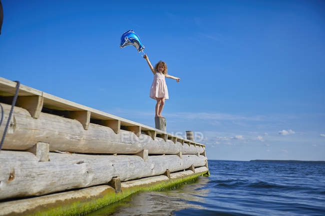 Junges Mädchen steht auf Holzpfosten auf Seebrücke und hält Heliumballon — Stockfoto