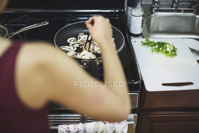 Jovem mulher mexendo cogumelos fatiados na frigideira, visão traseira — Fotografia de Stock