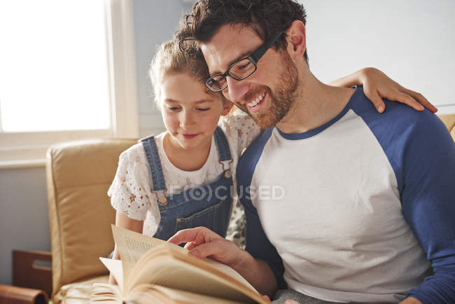 Hombre adulto medio leyendo libro con hija en sofá - foto de stock