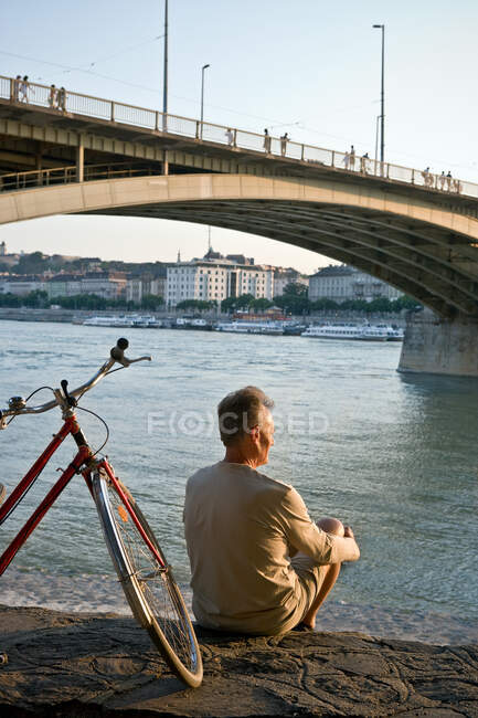 Hombre sentado solo junto al río con bicicleta - foto de stock