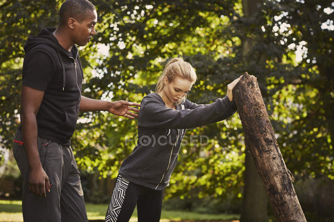Junge Frau mit Personal Trainer hebt Baumstamm in Park — Stockfoto
